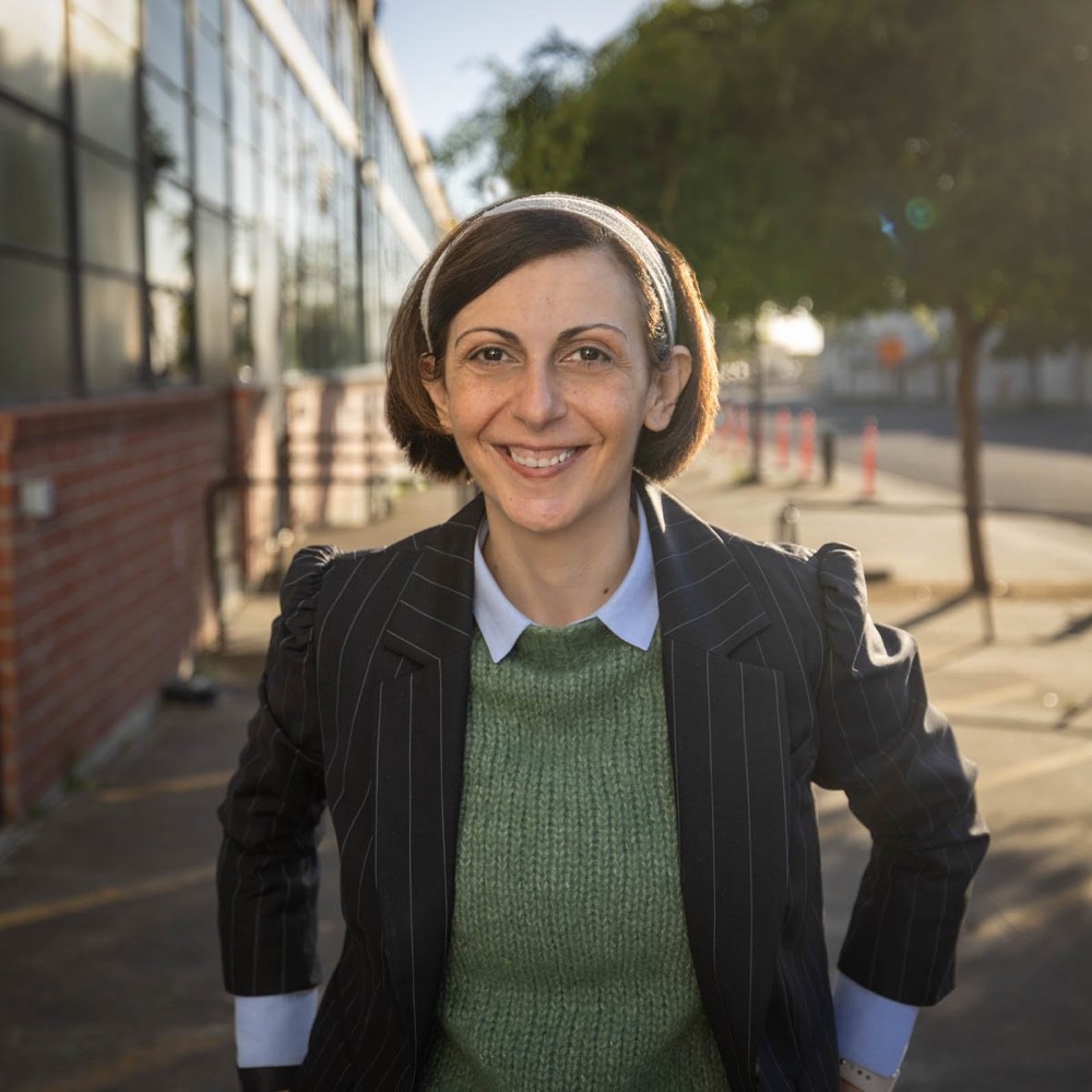 Annalisa Siagura standing on a San Francisco sidewalk in evening light, smiling, in a green sweater and pinstriped jacket